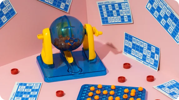 Gra bingo na różowym tle z niebiesko-żółtą maszyną do bingo, rozrzuconymi kulkami bingo, kartami i markerami, co wywołuje radosną atmosferę.